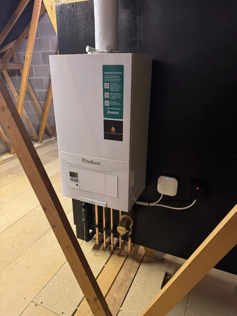 A wall-mounted boiler with HJT Heating Solutions branding and pipework in a loft